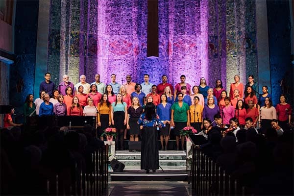 Members of the Tyndale Community Choir dressed in colourful shirts smiling on stage with a purple-lit background