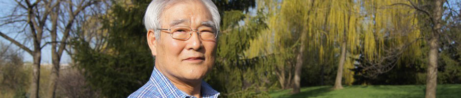 An older Korean man with glasses standing in the sunshine against a backdrop of green trees