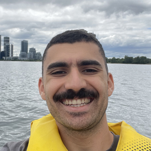 Dimo Ayoub in a life jacket on a lake. There is a city skyline behind him. 