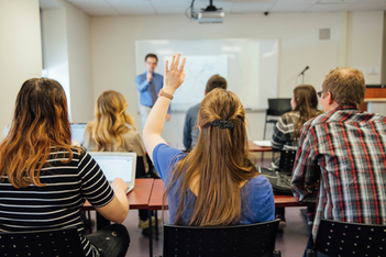 a class of students. A female student is raising her hand to ask the teacher a question.